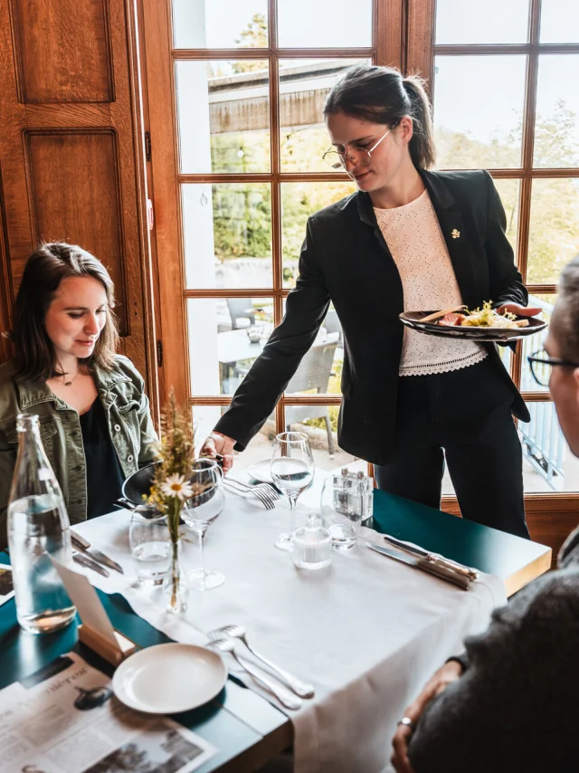 Un moment convivial dans un restaurant élégant et chaleureux. Une serveuse élégamment vêtue sert un plat raffiné à une table où un homme et une femme profitent d'un repas. La table est dressée avec soin, ornée de fleurs fraîches, de verres à pied et de menus. La lumière naturelle illumine la scène à travers de grandes fenêtres en bois, offrant une vue sur un jardin extérieur verdoyant.